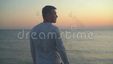 一个年轻人站在海上日落的背景上。 一个人背对着摄像机站着，站在海滩上。